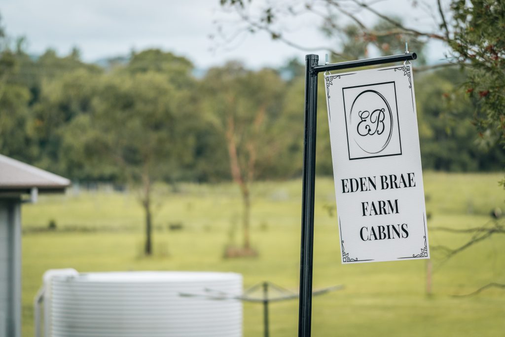 "Eden Brae Farm Cabins sign on a black post, set against a picturesque rural landscape with trees and a water tank in the background.